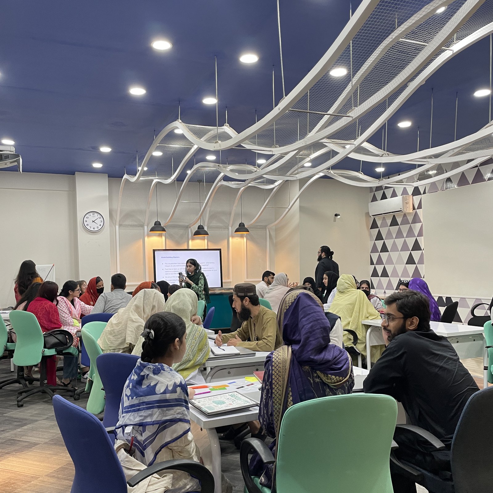 The full workshop room mid-session: a facilitator presenting World Building Starters on screen while teams work through cards and sticky notes at their tables.