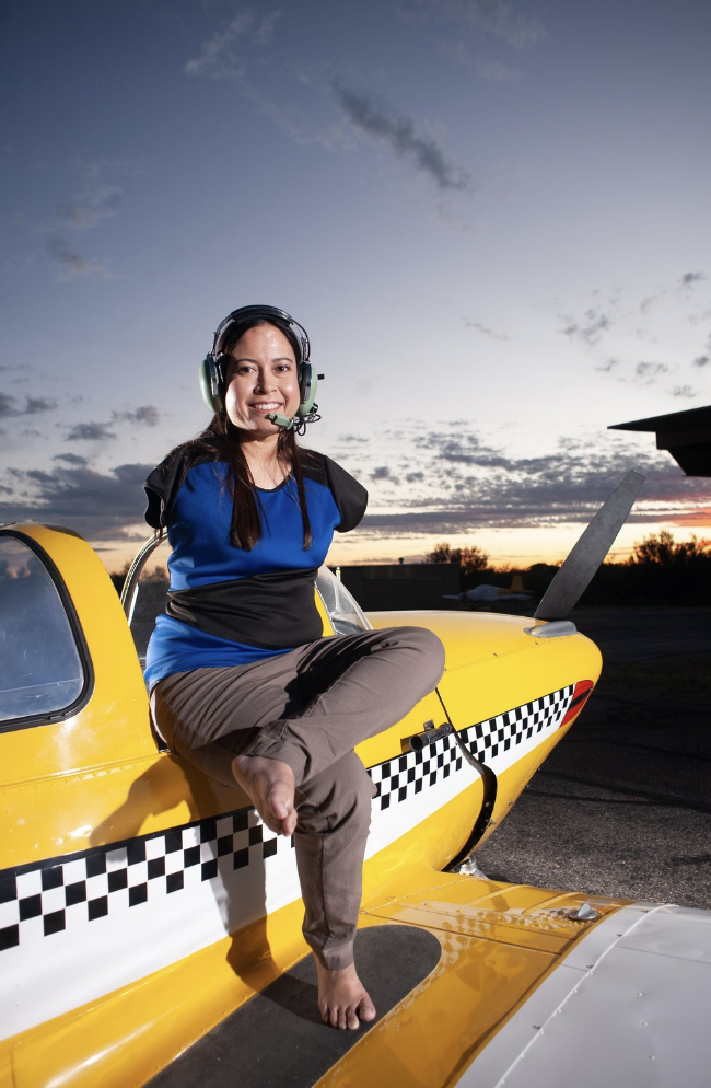 Jessica Cox, barefoot, seated in the open cockpit of a small yellow aircraft at dusk, wearing a pilot headset.